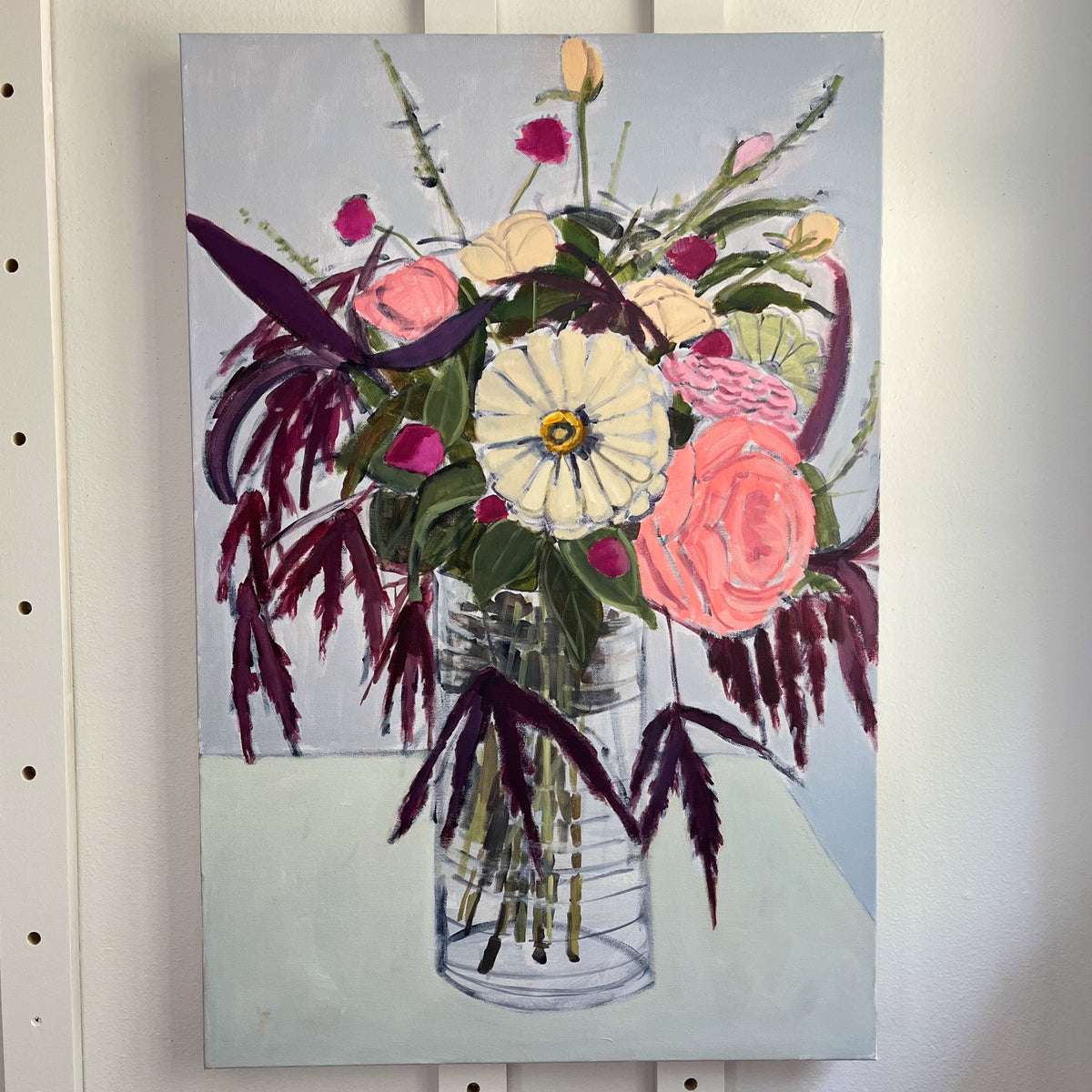 September Bouquet — a tall bouquet of pink, cream, and yellow flowers with dark trailing leaves in a clear vase. Painting by Maggie Stickney.