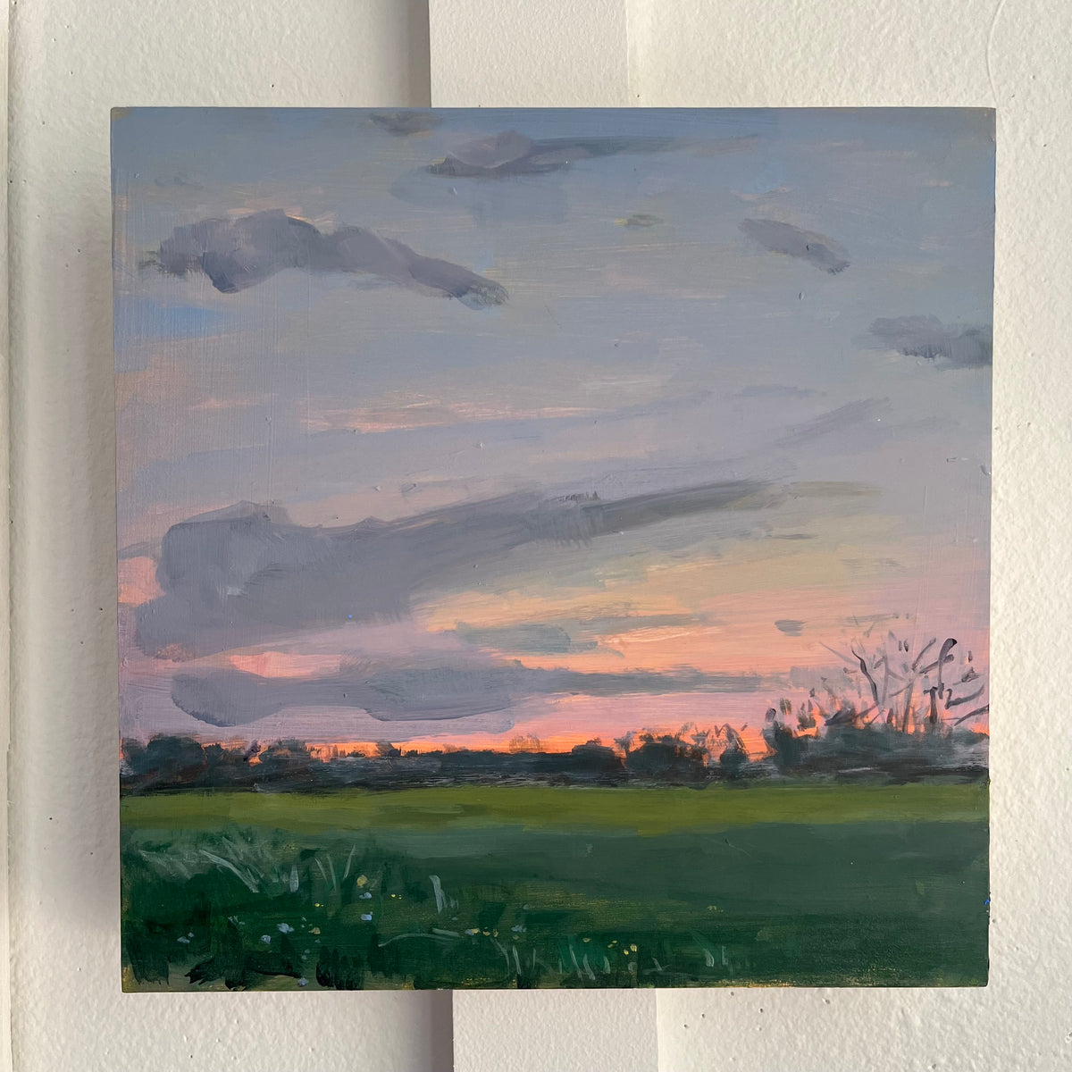 Last Light — a green field and dark tree line beneath streaks of pink sunset clouds. Painting by Maggie Stickney.