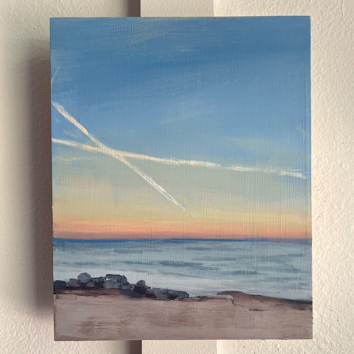Long Run no 2 — white streaks crossing a blue evening sky above calm water and a sandy shore. Painting by Maggie Stickney.
