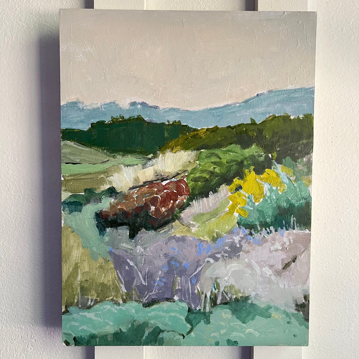 English Garden — layered garden beds in green, purple, and yellow with hills beyond. Painting by Maggie Stickney.